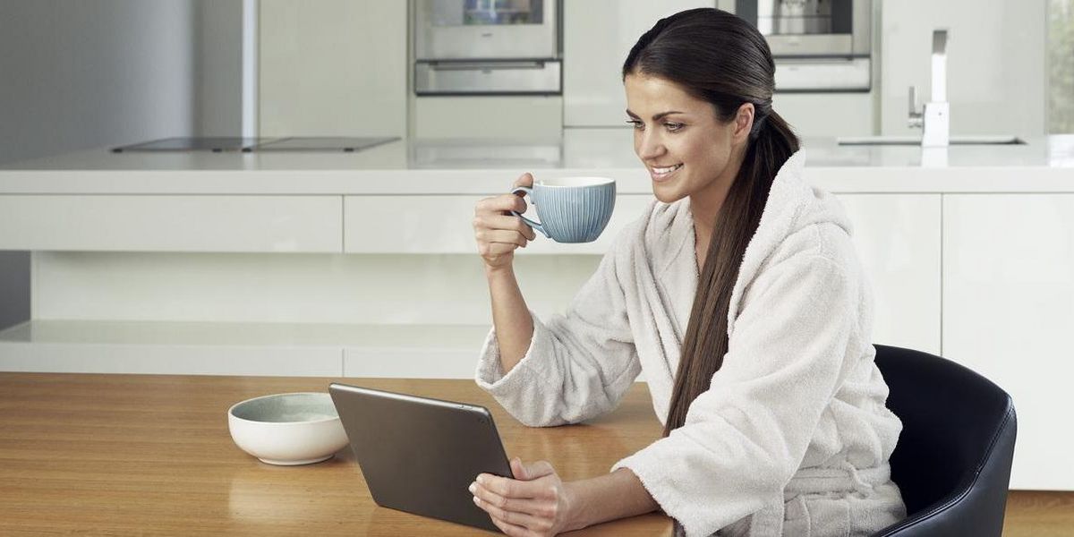 Eine Frau sitzt lächelnd mit einer Tasse in der Hand in der Küche vor einem Laptop.