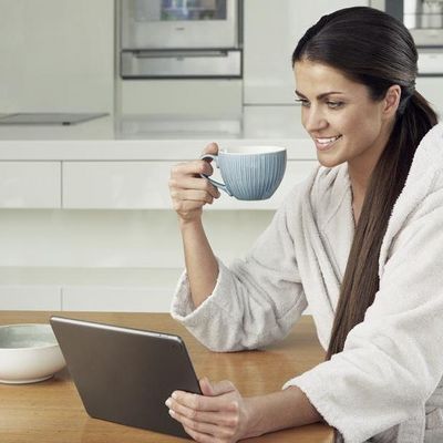 Eine Frau sitzt lächelnd mit einer Tasse in der Hand in der Küche vor einem Laptop.