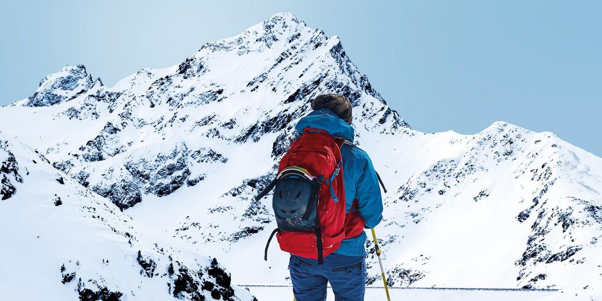 Eine Person steht im Freien und trägt einen Skianzug, einen Rucksack und Skistöcke. Sie schaut auf die schneebedeckten Berge.