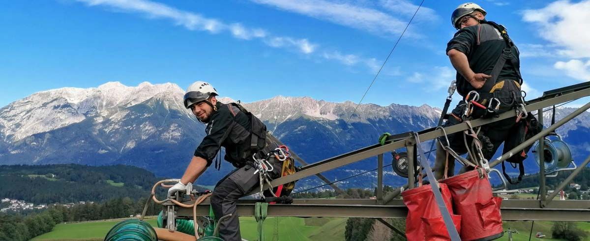 Zwei Männer sitzen mit Gurten und Helm auf einem Strommasten.
