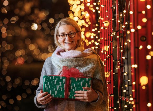 Eine fröhliche Frau hält ein eingepacktes Geschenk in der Hand, während sie im Freien steht, umgeben von festlicher Weihnachtsbeleuchtung. Sie verkörpert den fröhlichen Geist der Weihnachtszeit.