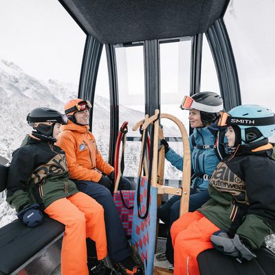 Vier Personen sitzen in einer Gondel mit Skianzug und Helm und haben Rodeln mit.