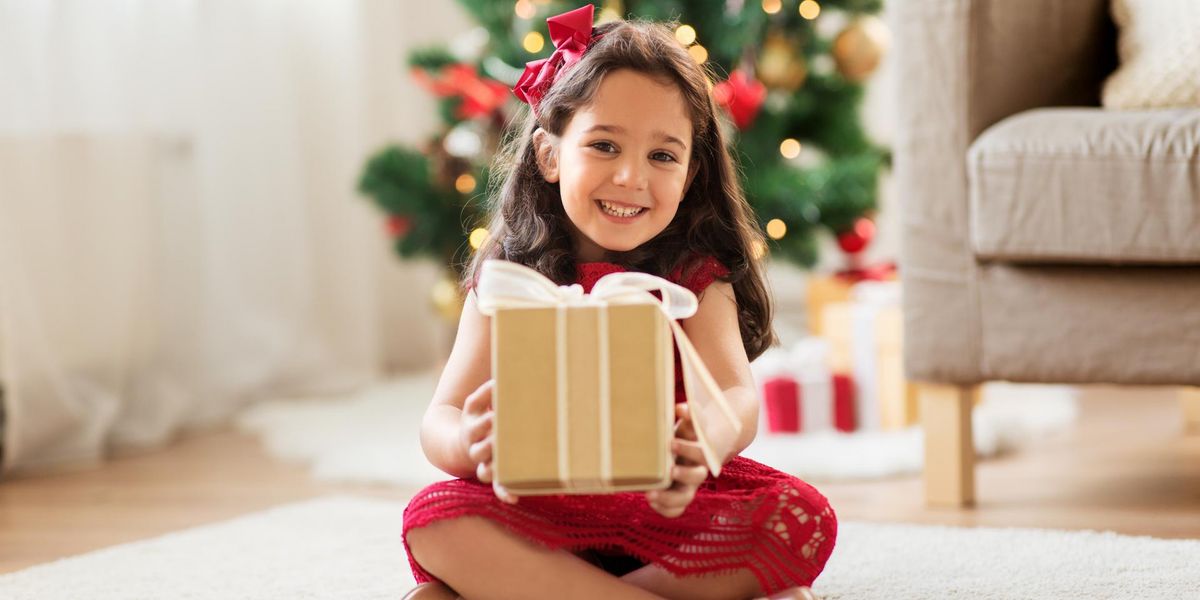 Ein lachendes Mädchen mit einem Geschenk in der Hand. Sie sitzt vor einem Weihnachtsbaum.