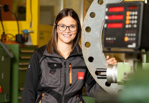 Eine Frau steht lachend in ihrer Arbeitskleidung neben einer Maschine.