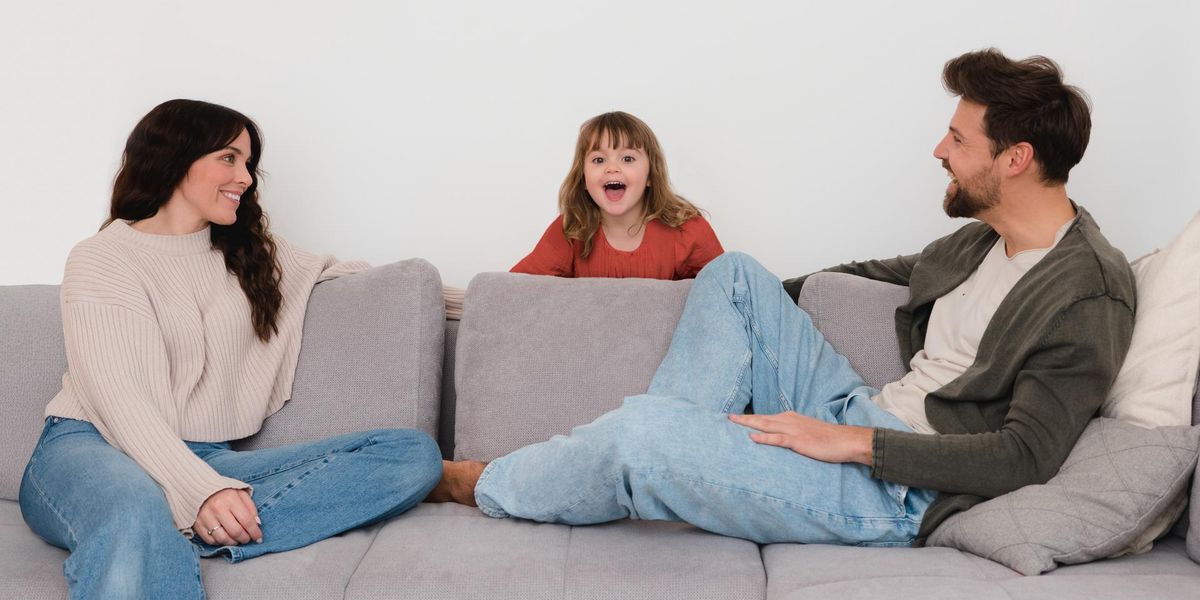 Mutter und Vater sitzen auf dem Sofa und schauen ihr Tochter, die zwischen ihnen hinter einem Polster sitzt, lachend an.