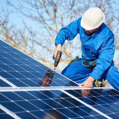 Männlicher Ingenieur in blauem Anzug und Schutzhelm bei der Installation von Photovoltaikmodulen mit einem Schraubenzieher. Professioneller Elektriker bei der Montage von Solarmodulen auf dem Dach. Ökologisches Konzept der alternativen Energie.