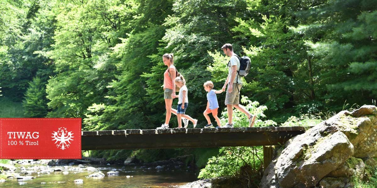 Eine Familie geht über eine Holzbrücke. Unter der Brücke fließt ein Fluss und rundherum sind Bäume und Steine.