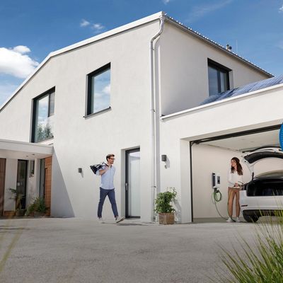 Ein Haus mit Garage, in der ein E-Auto parkt, das gerade aufgeladen wird. Die Dame steckt es ab und der Mann ist auf den Weg zum Auto.