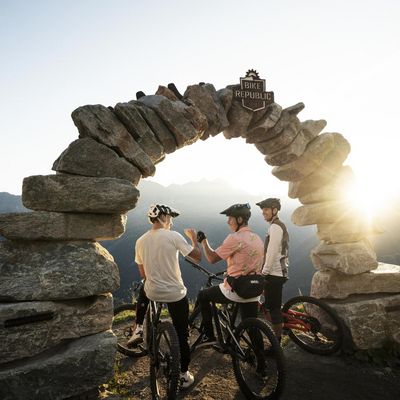 Drei Personen am Rad stehen unter einem Bogen aus Steinen und schlagen mit den Fäusten ein. Im Hintergrund geht die Sonne unter.